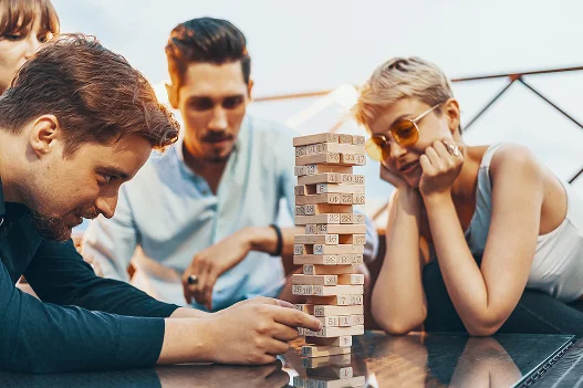 Vier mensen kijken gespannen naar een Jenga-toren op een glazen tafel. Eén van hen trekt voorzichtig een blokje eruit. De sfeer is gespannen, maar toch speels.