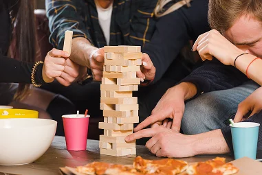 Een groep mensen speelt geconcentreerd Jenga en kijkt aandachtig naar een toren van houten blokken. Op de tafel staan bekers en kommen, met op de voorgrond een pizza.